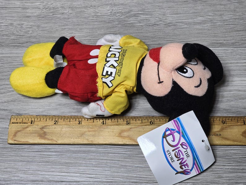 K&ouml;nnte beinhalten: Ein gelbes, rotes und schwarzes Pl&uuml;sch-Mickey-Mouse-Spielzeug. Das Spielzeug tr&auml;gt ein gelbes Shirt mit dem Text "Disney Store" und "Mickey" darauf. Das Spielzeug liegt auf einer Holzoberfl&auml;che.