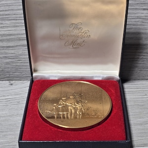 May include: A circular gold-toned commemorative medal featuring a relief of a family group, housed in a dark blue presentation box with a red velvet insert. The interior lid is lined with white satin and displays the text The Franklin Mint.
