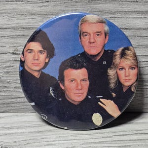 Puede incluir: Un botón redondo azul con cuatro personas en uniformes oscuros sobre un fondo azul. El botón parece ser un objeto de colección vintage, posiblemente relacionado con una serie de televisión o una película.