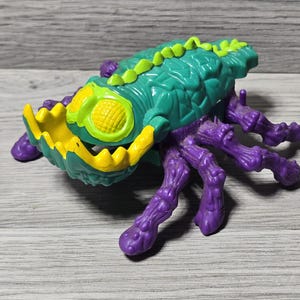 May include: A colorful toy insect with a textured teal body, yellow mouth, and purple legs. The insect has a segmented body and a row of green spikes along its back. The toy is made of plastic and sits on a gray wooden surface.