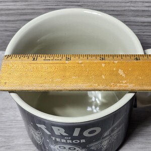 Puede incluir: Una taza de cer&aacute;mica blanca con una banda gris oscuro con el texto "TRIO OF TERROR" e ilustraciones de dibujos animados. Una regla de madera se coloca sobre la parte superior de la taza, midiendo aproximadamente 10 cm de ancho.