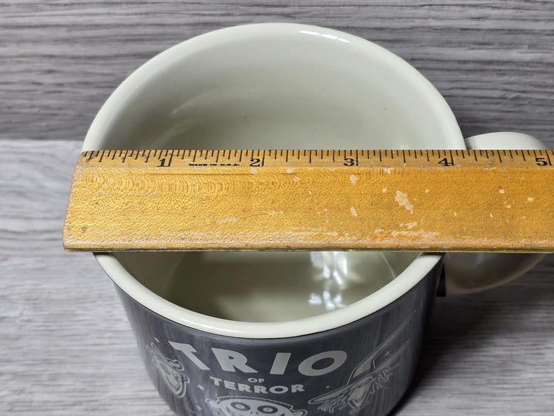 Puede incluir: Una taza de cer&aacute;mica blanca con una banda gris oscuro con el texto "TRIO OF TERROR" e ilustraciones de dibujos animados. Una regla de madera se coloca sobre la parte superior de la taza, midiendo aproximadamente 10 cm de ancho.
