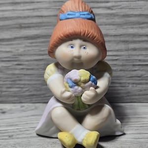 May include: A ceramic figurine of a seated girl with red hair and a blue bow. She wears a light pink dress with yellow accents and holds a bouquet of flowers. The figurine has blue eyes and freckles.