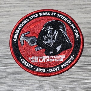 以下が含まれることがあります： ダース・ベイダーとスター・ウォーズをテーマにした、赤と黒の丸いパッチ。パッチには「GENERATIONS STAR WARS ET SCIENCE-FICTION」と「LES HERITIERS DE LA FORDE」の文字が含まれています。「2013」の年も表示されています。