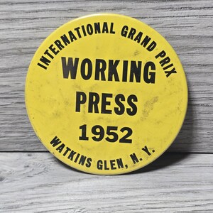 May include: A vintage yellow button with black text. The button reads "INTERNATIONAL GRAND PRIX WORKING PRESS 1952 WATKINS GLEN, N.Y." The button is circular and has a slightly worn appearance.