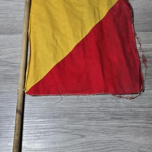 Puede incluir: Una bandera de señales náuticas vintage con un mástil de madera. La bandera está dividida diagonalmente, con una sección superior amarilla y una sección inferior roja. La bandera de tela muestra signos de desgaste.