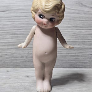 May include: A vintage porcelain doll with a blonde, curly hairstyle and delicate facial features, including rosy cheeks and large eyes. The doll has outstretched arms and is nude, showcasing its detailed craftsmanship. The doll is standing on a light-colored surface.