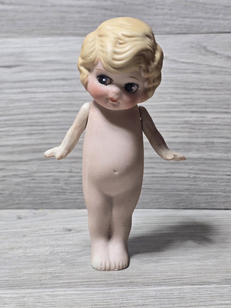 May include: A vintage porcelain doll with a blonde, curly hairstyle and delicate facial features, including rosy cheeks and large eyes. The doll has outstretched arms and is nude, showcasing its detailed craftsmanship. The doll is standing on a light-colored surface.