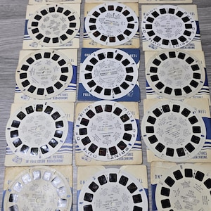 Könnte beinhalten: Eine Sammlung von Vintage-View-Master-Rollen und ihrer Verpackung. Die Rollen sind kreisförmig mit kleinen rechteckigen Sichtfenstern und sind weiß mit schwarzen Details. Die Verpackung ist hellbraun mit blauen Akzenten und Text.