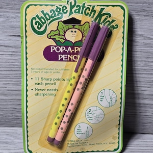 Może przedstawiać: Zestaw ołówków Pop-A-Point Cabbage Patch Kids w oryginalnym opakowaniu. Ołówki są żółte i różowe z fioletowymi akcentami i kwiatowym wzorem. Opakowanie jest zielono-żółte z logo Cabbage Patch Kids.