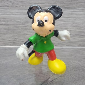 Pode incluir: Uma figurinha de pl&aacute;stico vintage do Mickey Mouse vestindo uma camisa verde, cal&ccedil;as vermelhas e sapatos amarelos. A figurinha est&aacute; de p&eacute; com os bra&ccedil;os estendidos.