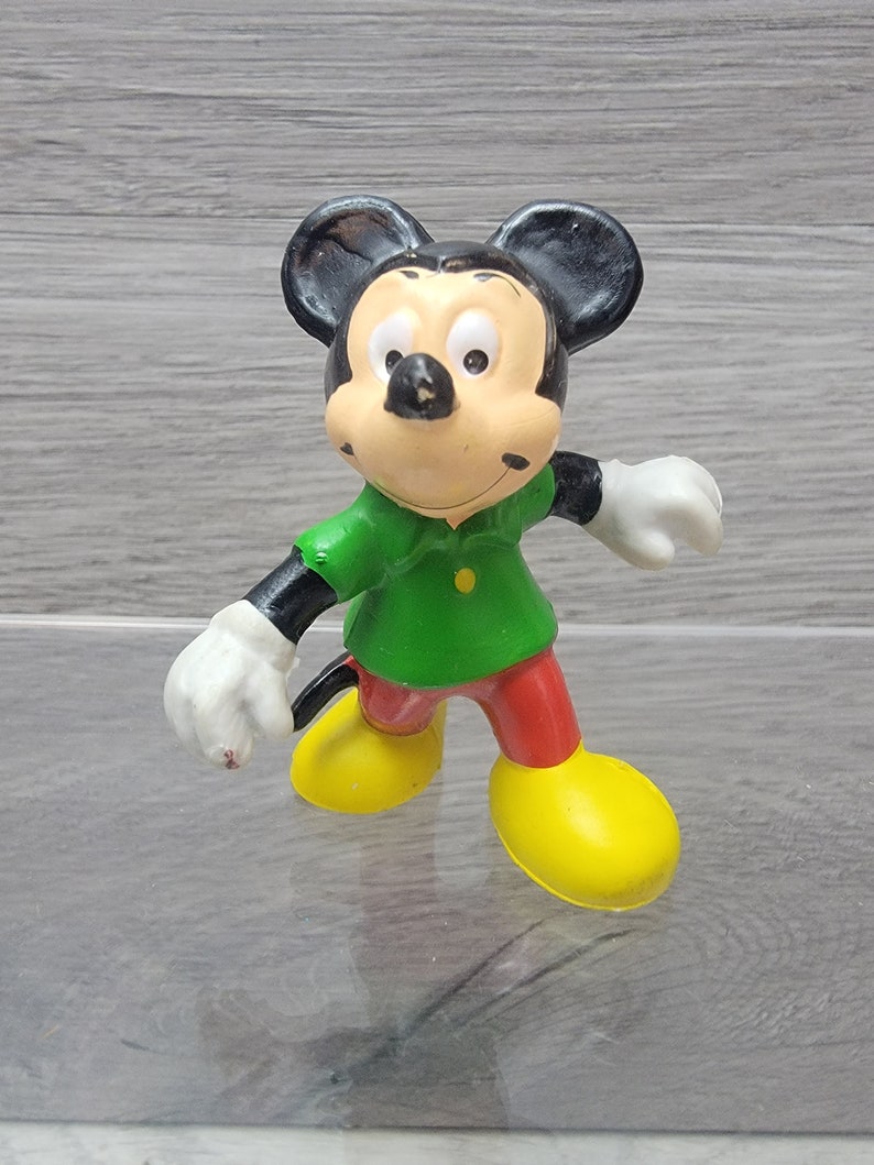 Pode incluir: Uma figurinha de pl&aacute;stico vintage do Mickey Mouse vestindo uma camisa verde, cal&ccedil;as vermelhas e sapatos amarelos. A figurinha est&aacute; de p&eacute; com os bra&ccedil;os estendidos.