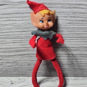Puede incluir: Una figura de elfo vintage con un atuendo, sombrero y piernas de fieltro rojo. El elfo tiene una cara de plástico con ojos azules, una nariz pequeña y cabello amarillo. Un cuello de fieltro gris completa el atuendo. La figura está de pie.