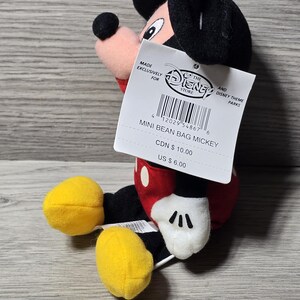 以下が含まれることがあります： 小さなミッキーマウスのビーンバッグぬいぐるみ。ぬいぐるみは、黒い頭と耳、赤い体、黒いディテールのある白い手袋、黄色の足を持っています。「MINI BEAN BAG MICKEY」と書かれたタグが付いています。