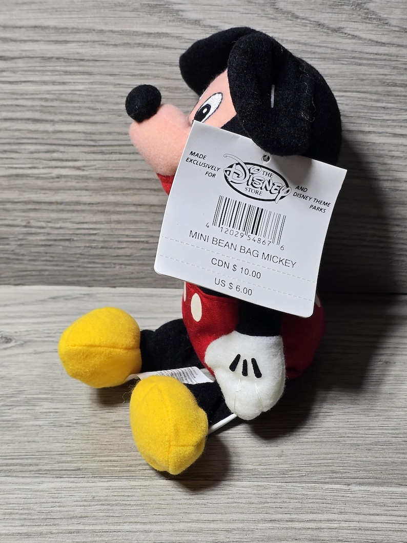 以下が含まれることがあります： 小さなミッキーマウスのビーンバッグぬいぐるみ。ぬいぐるみは、黒い頭と耳、赤い体、黒いディテールのある白い手袋、黄色の足を持っています。「MINI BEAN BAG MICKEY」と書かれたタグが付いています。