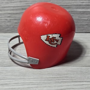 Op de afbeelding: Een rode plastic American footballhelm met een grijs gezichtsmasker. De helm heeft een wit en rood logo aan de zijkant. Een sport verzamelobject.