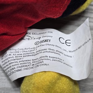 K&ouml;nnte beinhalten: Ein wei&szlig;es Etikett mit schwarzem Text, das an einem roten und gelben Pl&uuml;schtier befestigt ist. Das Etikett lautet "MADE EXCLUSIVELY FOR DISNEY Company DISNEY The Disney Store, Ltd., Watford WDI 8BX.U.K. CE PA1082/Ohio 12441/ 7-537-M. All new materials, polyester fibre, plastic pellets/ Materiels neuf, fibres synthetiques et cellulosiques, new material, polyester faser, plastik kugelchen/ pastilles en plastique/ amente nuevo material, fibra polyester, pelottila plastico MADE IN CHINA/FABRIKAT IN CHINA  MADE IN CHINE/HECHO EN CHINA"