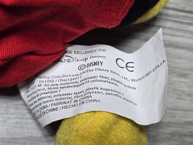 K&ouml;nnte beinhalten: Ein wei&szlig;es Etikett mit schwarzem Text, das an einem roten und gelben Pl&uuml;schtier befestigt ist. Das Etikett lautet "MADE EXCLUSIVELY FOR DISNEY Company DISNEY The Disney Store, Ltd., Watford WDI 8BX.U.K. CE PA1082/Ohio 12441/ 7-537-M. All new materials, polyester fibre, plastic pellets/ Materiels neuf, fibres synthetiques et cellulosiques, new material, polyester faser, plastik kugelchen/ pastilles en plastique/ amente nuevo material, fibra polyester, pelottila plastico MADE IN CHINA/FABRIKAT IN CHINA  MADE IN CHINE/HECHO EN CHINA"