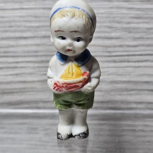 Pu&ograve; includere: Statuina vintage in porcellana di un ragazzino vestito da marinaio. La statuina indossa un cappello da marinaio bianco con una striscia blu, una camicia bianca con colletto blu e pantaloncini verdi. Tiene in mano una piccola barca a vela gialla e rossa.