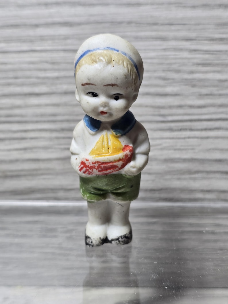 Pu&ograve; includere: Statuina vintage in porcellana di un ragazzino vestito da marinaio. La statuina indossa un cappello da marinaio bianco con una striscia blu, una camicia bianca con colletto blu e pantaloncini verdi. Tiene in mano una piccola barca a vela gialla e rossa.