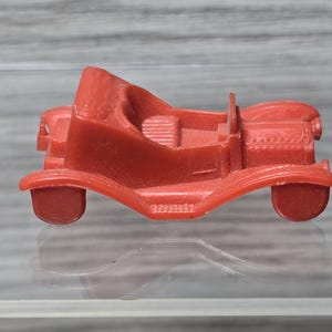 Puede incluir: Un coche de juguete de plástico rojo con un diseño detallado. El coche tiene una carrocería redondeada y un diseño simple de techo abierto.