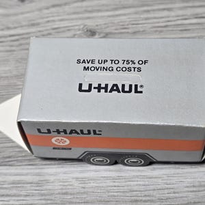 May include: A silver U-Haul box with the text "SAVE UP TO 75% OF MOVING COSTS" and the U-Haul logo. The box has an orange stripe and a graphic of a trailer on the side. The box is on a wooden surface.