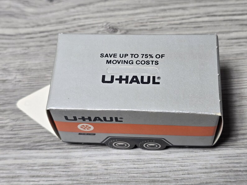May include: A silver U-Haul box with the text "SAVE UP TO 75% OF MOVING COSTS" and the U-Haul logo. The box has an orange stripe and a graphic of a trailer on the side. The box is on a wooden surface.