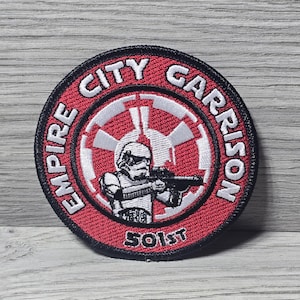 以下が含まれることがあります： 赤色の背景と黒い縁取りの丸いパッチ。パッチには、白い文字で「EMPIRE CITY GARRISON」と「501st」と書かれています。ストームトルーパーの白黒イラストが中央にあります。