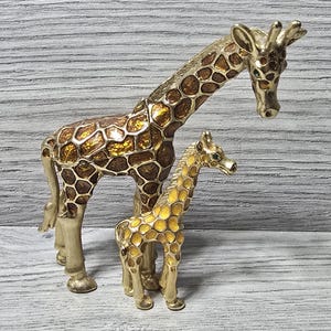 May include: Two decorative giraffe figurines. The larger giraffe has a brown and gold patterned body with gold legs and a long neck. The smaller giraffe has a gold patterned body and gold legs. Both have green eyes.