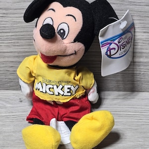 K&ouml;nnte beinhalten: Ein gelber Pl&uuml;sch-Mickey Mouse, der ein gelbes Shirt mit dem Text "The Spirit of Mickey" und rote Shorts tr&auml;gt. Der Pl&uuml;sch sitzt auf einer Holzoberfl&auml;che mit einem wei&szlig;en Etikett, auf dem "The Disney Store" steht.