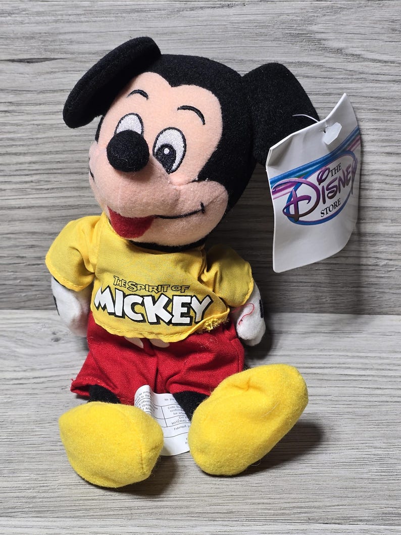 K&ouml;nnte beinhalten: Ein gelber Pl&uuml;sch-Mickey Mouse, der ein gelbes Shirt mit dem Text "The Spirit of Mickey" und rote Shorts tr&auml;gt. Der Pl&uuml;sch sitzt auf einer Holzoberfl&auml;che mit einem wei&szlig;en Etikett, auf dem "The Disney Store" steht.