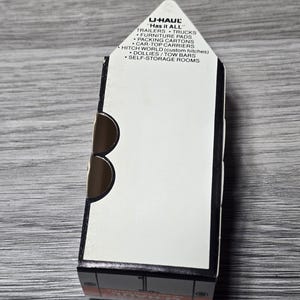 May include: A U-Haul box featuring the text "Has it ALL" and a list of services such as trailers, trucks, and self-storage rooms. The box is white with black and brown details, resting on a grey wooden surface.