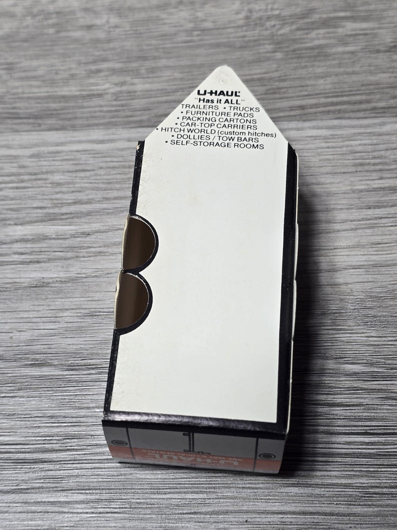 May include: A U-Haul box featuring the text "Has it ALL" and a list of services such as trailers, trucks, and self-storage rooms. The box is white with black and brown details, resting on a grey wooden surface.