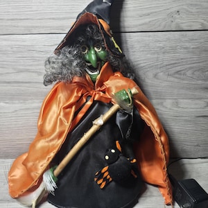 May include: A witch figurine with a green face, grey hair, and a black pointed hat featuring yellow stars. The figure wears a shiny orange cape over a black dress, holds a wooden broomstick, and has a plush black spider with orange legs attached.