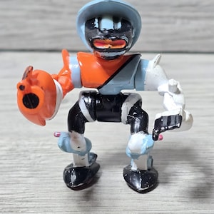 May include: A small, articulated robot toy with a light blue helmet and arms, an orange chest, and black legs. The robot has a black face with red details and an orange hand with a black circle. The toy is made of plastic.