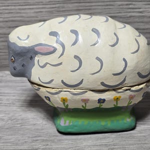 Peut inclure: Une figurine de mouton en céramique blanche avec des traits faciaux gris et un motif floral peint autour de la base. Le mouton est une boîte à charnières avec un couvercle amovible.