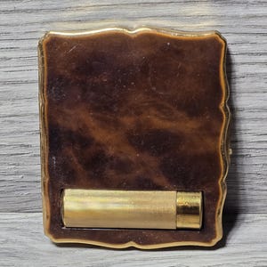 Può includere: Un astuccio compatto rettangolare con una finitura marmorizzata marrone scuro simile al tartarugato e un bordo smerlato in metallo dorato. Sulla parte anteriore è fissato un porta rossetto cilindrico orizzontale in metallo dorato con una sottile trama a coste.