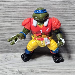 以下が含まれることがあります： アメフトのユニフォームを着たミュータント・タートルズのアクションフィギュア。フィギュアは緑色で、青いマスク、赤い肩パッド、黄色のズボンをしています。胸には1番が書かれており、黒い靴を履いています。
