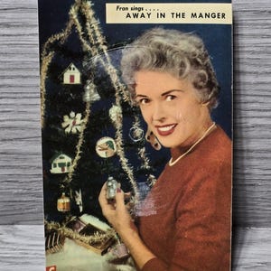May include: A vintage Christmas card featuring a woman in a red sweater holding a small silver bell ornament. The card has a blue background with a Christmas tree decorated with ornaments and tinsel. The text on the card reads "Fran sings... AWAY IN THE MANGER".