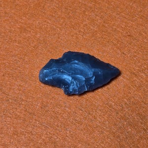 May include: A dark blue, irregularly shaped stone with a rough, textured surface. The stone is set against a textured orange background, creating a contrast in color and texture. The object appears to be a natural, possibly hand-worked, artifact.