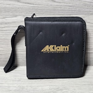 May include: Black zippered case with a handle. The case has the gold text "AKLAIM entertainment, inc." on the front. The case is square and has a black zipper and a black handle.