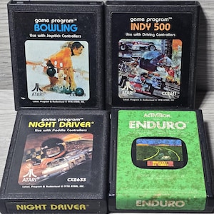 May include: Four vintage Atari game cartridges: Bowling, Indy 500, Night Driver, and Enduro. The Bowling cartridge features a bowler graphic, while Indy 500 shows race cars. Night Driver has a race car image, and Enduro displays a road scene. All are black cartridges.