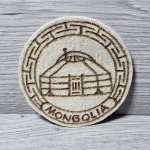May include: Round, beige felt coaster with a detailed, burnt-wood design. The design features a yurt, a traditional Mongolian dwelling, encircled by a geometric border. The word "MONGOLIA" is written along the bottom edge.