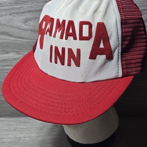May include: A vintage Ramada Inn trucker hat. The hat has a red peak and a white front panel with the words "RAMADA INN" in red block letters. The sides and back are red mesh.
