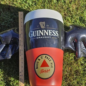 May include: A black and red inflatable Guinness pint glass with wings. The glass has the Guinness logo and the text "Draught". The bottom of the glass has a Bass Pale Ale logo.