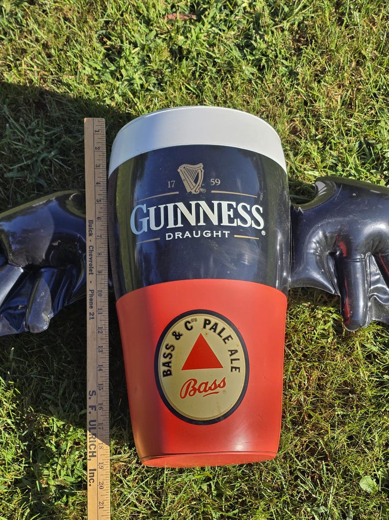May include: A black and red inflatable Guinness pint glass with wings. The glass has the Guinness logo and the text "Draught". The bottom of the glass has a Bass Pale Ale logo.
