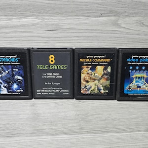 Könnte beinhalten: Vier schwarze, rechteckige Atari-Spielkassetten, die horizontal nebeneinander liegen. Die Etiketten zeigen die Titel Asteroids, 8 Tele-Games, Missile Command und Video Pinball sowie die dazugehörigen grafischen Illustrationen und Programmtext.