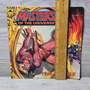 Puede incluir: Un c&oacute;mic vintage de "Masters of the Universe" con un personaje musculoso colgado de una esfera de reloj. La portada presenta el t&iacute;tulo en rojo y azul, con la palabra "TIME" visible. Una regla de madera est&aacute; colocada junto al libro.