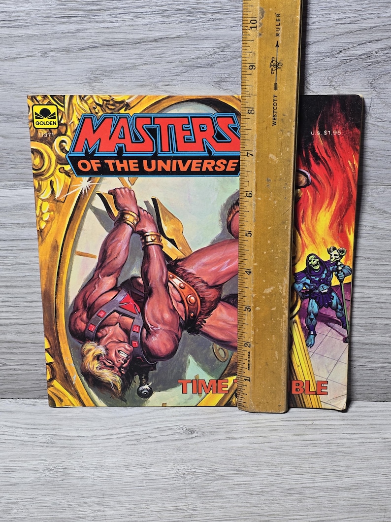 Puede incluir: Un c&oacute;mic vintage de "Masters of the Universe" con un personaje musculoso colgado de una esfera de reloj. La portada presenta el t&iacute;tulo en rojo y azul, con la palabra "TIME" visible. Una regla de madera est&aacute; colocada junto al libro.