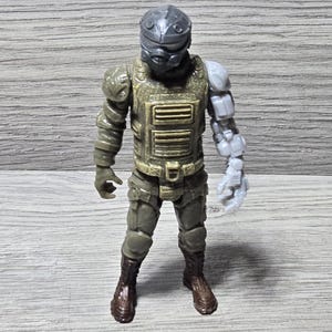 Peut inclure: Une figurine articulée en plastique représentant un soldat portant un casque gris foncé, un gilet tactique vert olive texturé, un pantalon assorti et des bottes marron. La figurine possède un bras gauche robotique gris clair distinct.